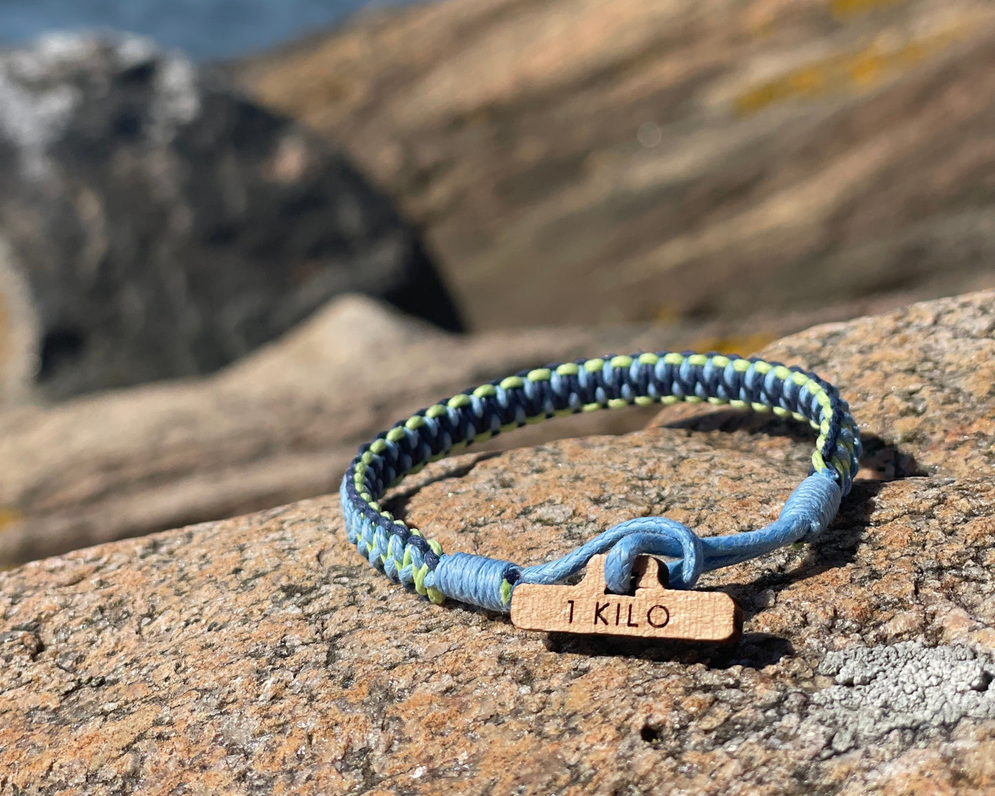 Blue and green braided bracelet with a wooden tag labeled '1 KILO' on a rocky surface.