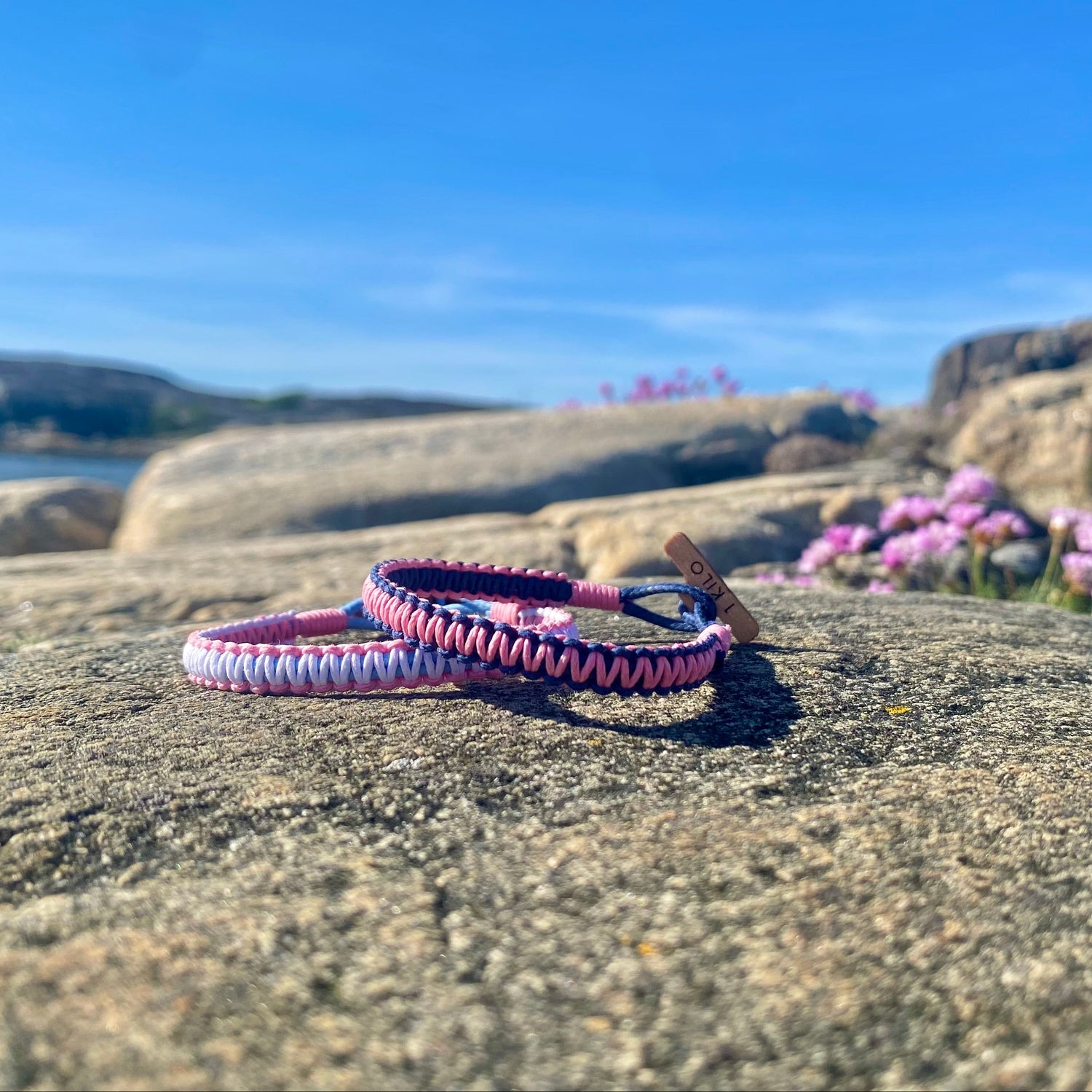 Two pink and blue braided bracelets on a rock with a scenic background of water, flowers and cliffs.